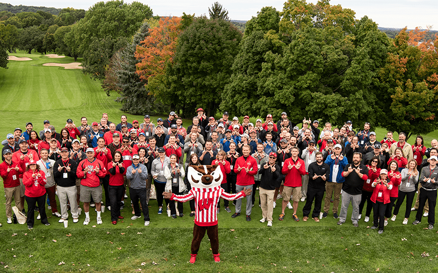 A large group of golfers and Bucky Badger pose for a group shot at the golf course.