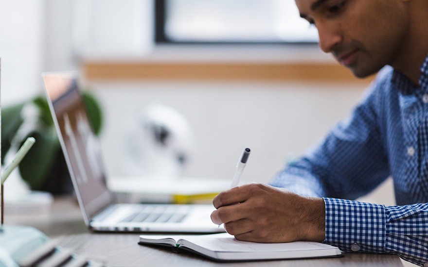 A man seating at a desk writing with a laptop next to him