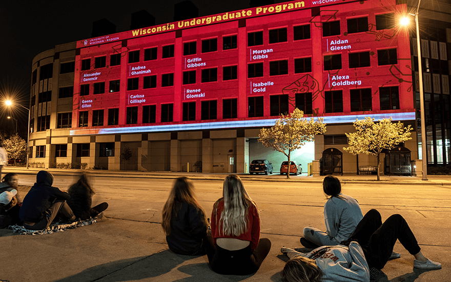 Passersby watch as the exterior of Grainger Hall is illuminated with congratulatory messages for graduates