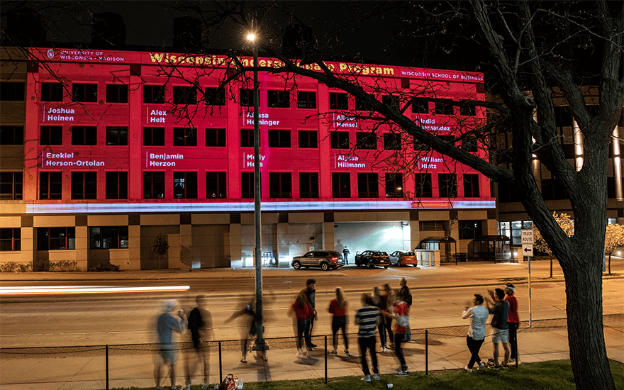 Passersby watch as the exterior of Grainger Hall is illuminated with congratulatory messages for graduates