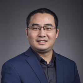 Professor Cheng He portrait, man with dark hair smiling, wearing a navy blue jacket and black button up shirt.