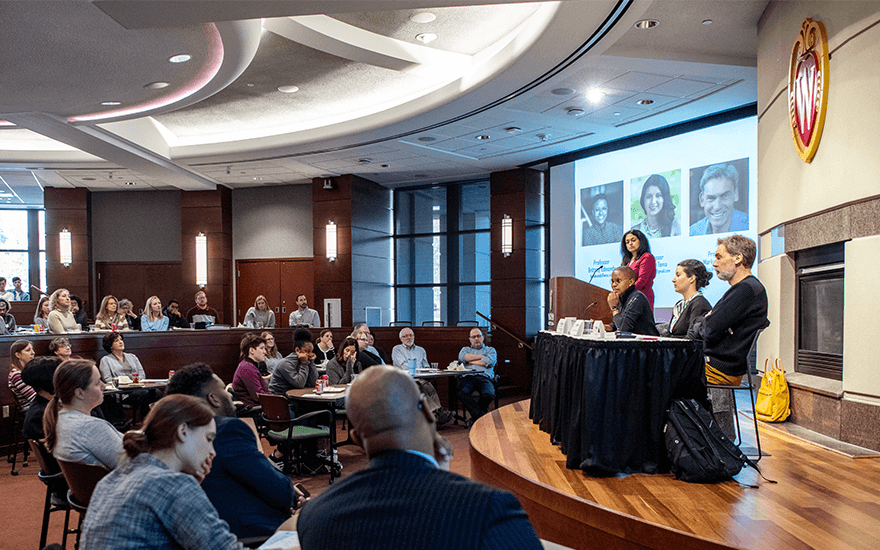 A large group of faculty, staff, and students listen to a panel of speakers