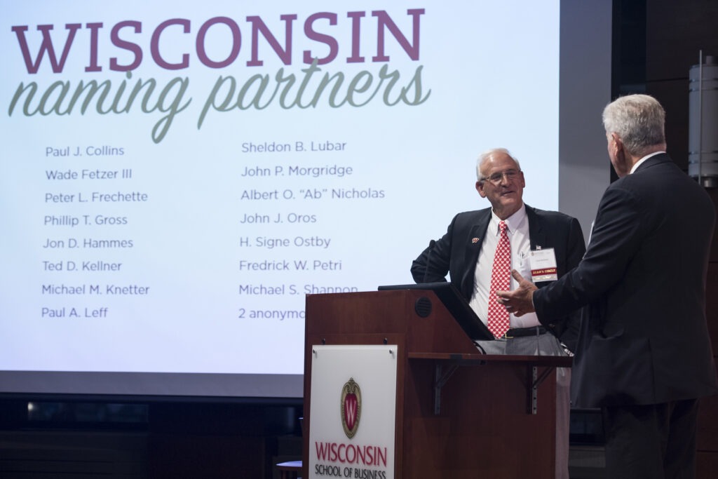 Two men on a stage with Wisconsin naming partners on the screen behind them.