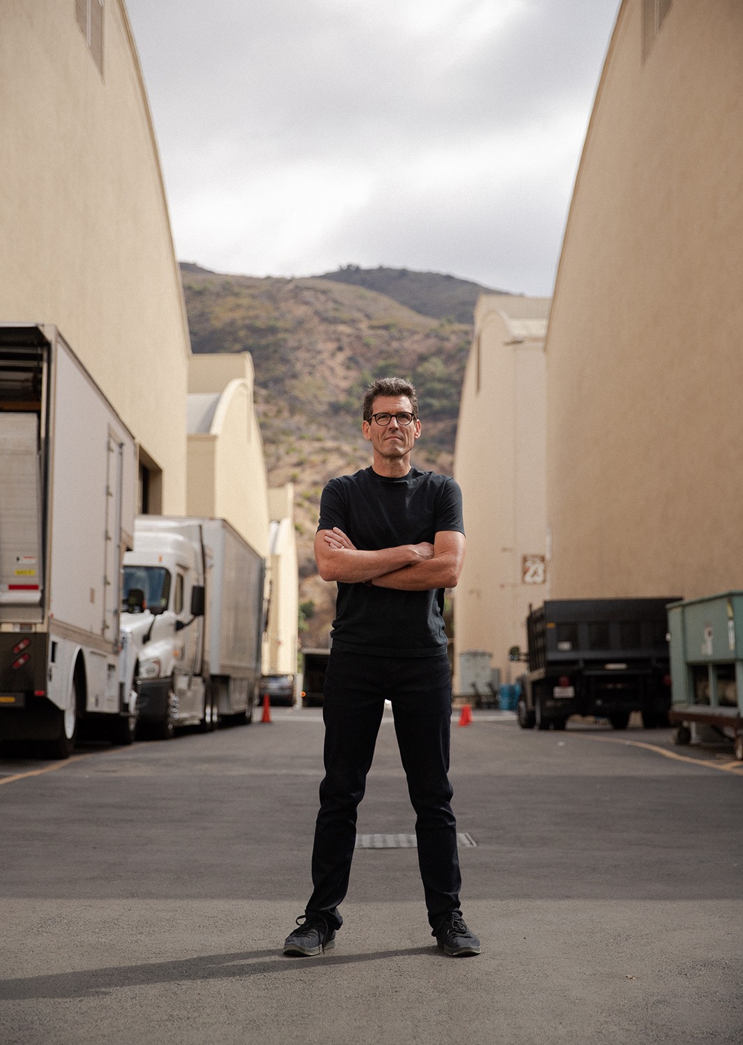 Jim standing with his arms crossed in between several buildings and parked trucks