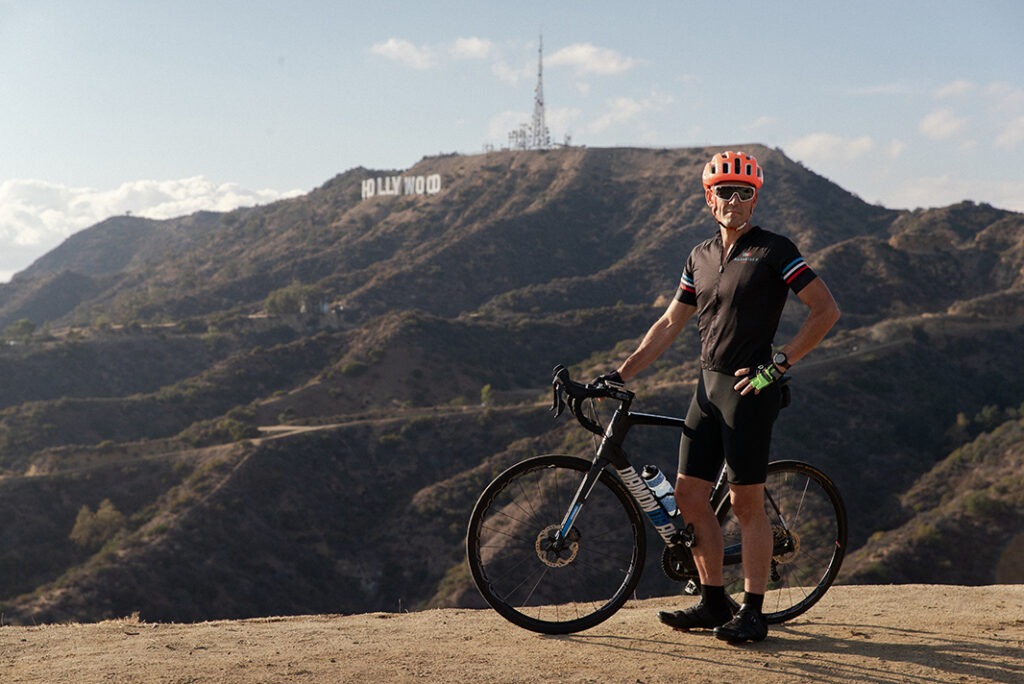 Jim in a bike helmet and sunglasses, posing with his bike in front of the Mount Lee and the  Hollywood sign