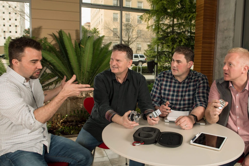 Four men sitting around a table with the man on the left talking and making gestures with his hands