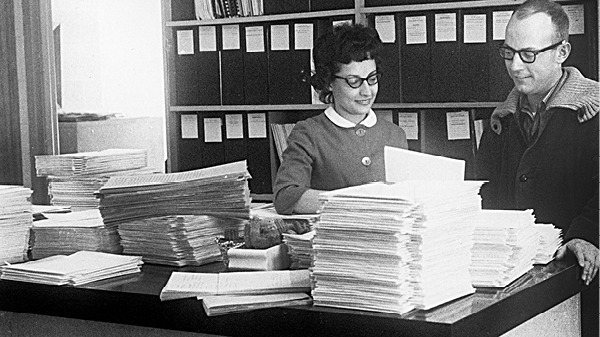 A man and a woman standing in front of a desk looking at records
