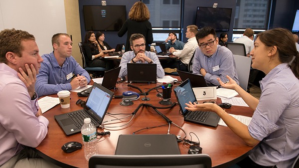 Five students sitting at a round table with laptops open in front of them are in a discussion