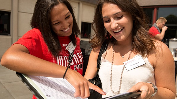 One female student is holding a folder and another female student is pointing at the notes in the folder