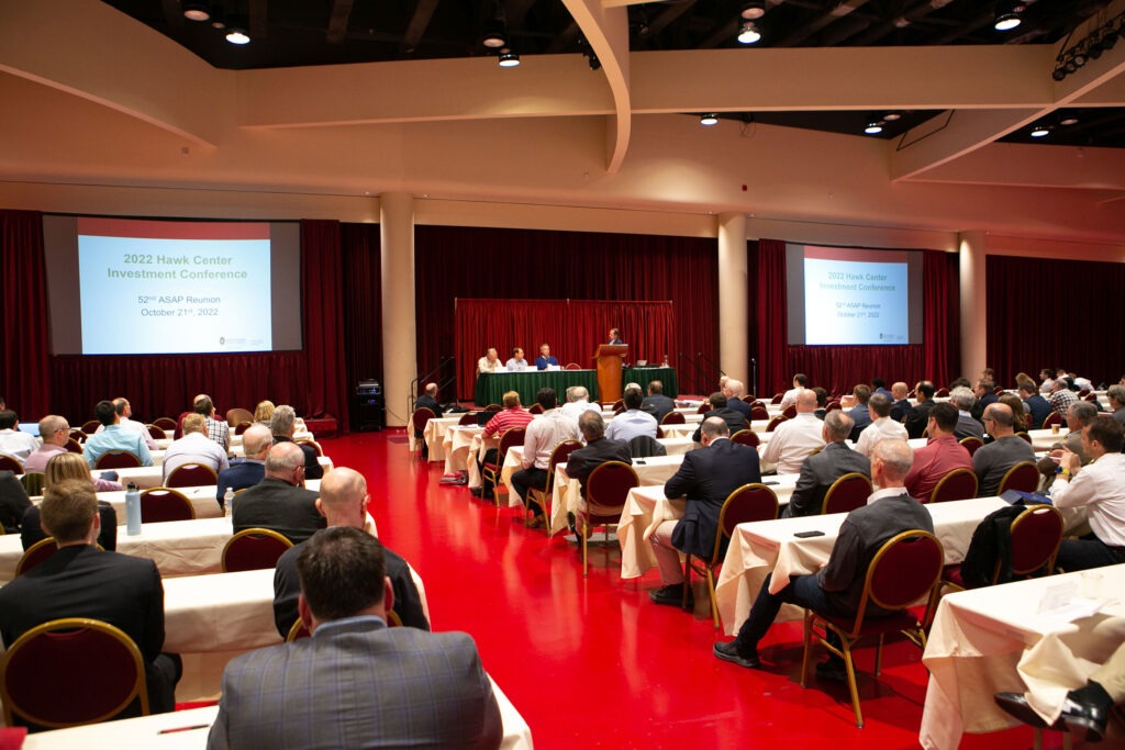 A group of people sitting in chairs facing a group of three people siting and one man talking at a podium at the 2022 Hawk Center Investment Conference