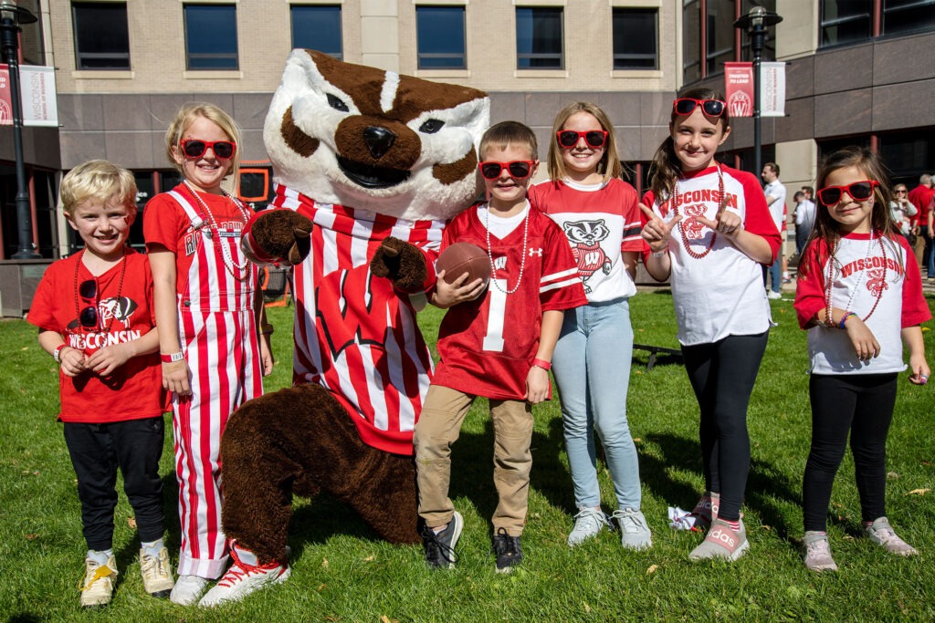 A group of children clad in red and white pose with Bucky Badger outside Grainger Hall.