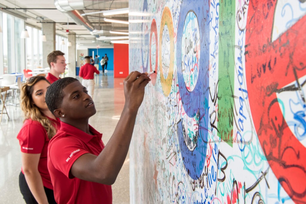 Three students wearing red BEL Program shirts are writing on a wall that is covered with writing.