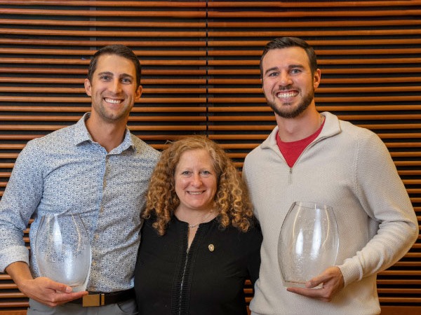 Tyler Kennedy and Wes Schroll holding large glass containers with a woman standing between them.
