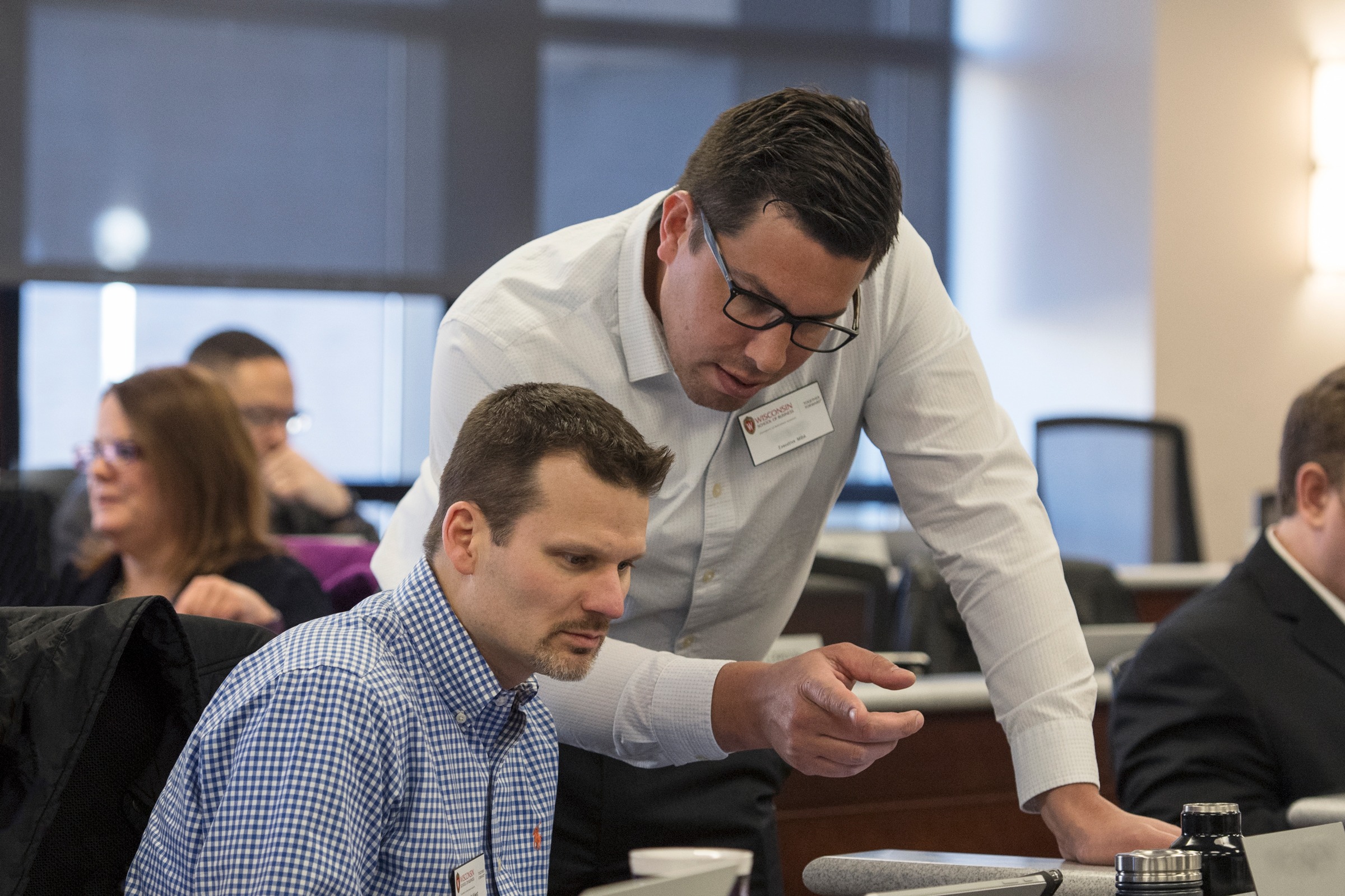 One man sitting and another one standing near him while pointing at a tablet