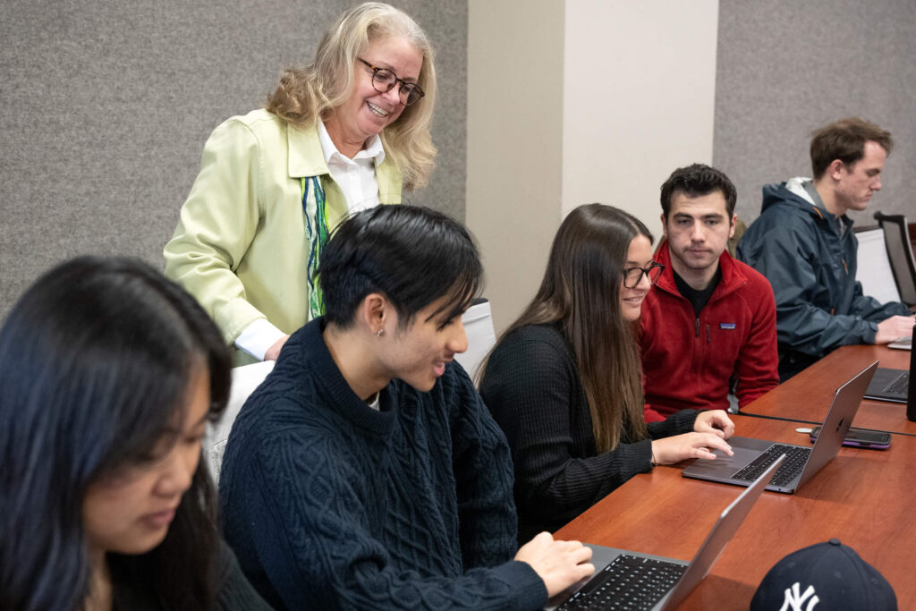 A woman standing with five students sitting at a desk with their laptops in front of them.