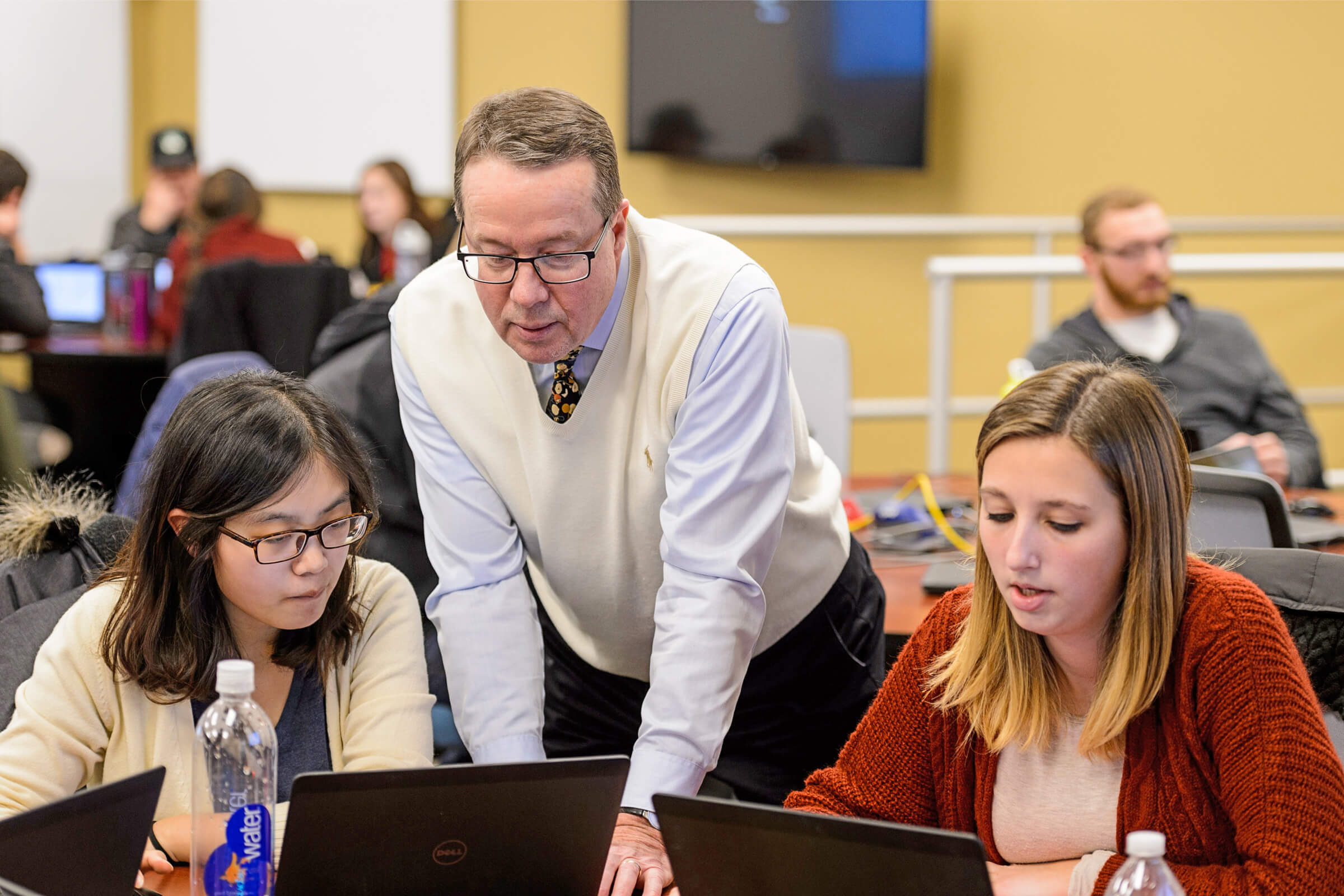 A man leaning on a desk between two women sitting at a desk looking at their laptops in front of them.