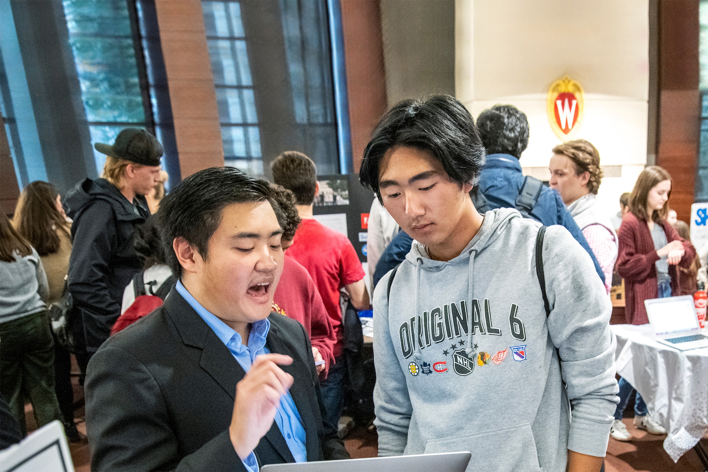 Two men in a discussion while looking at a laptop screen with students walking around behind them.