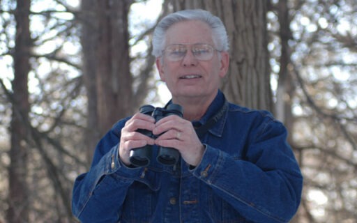 Eisele smiling as he holds binoculars in the woods