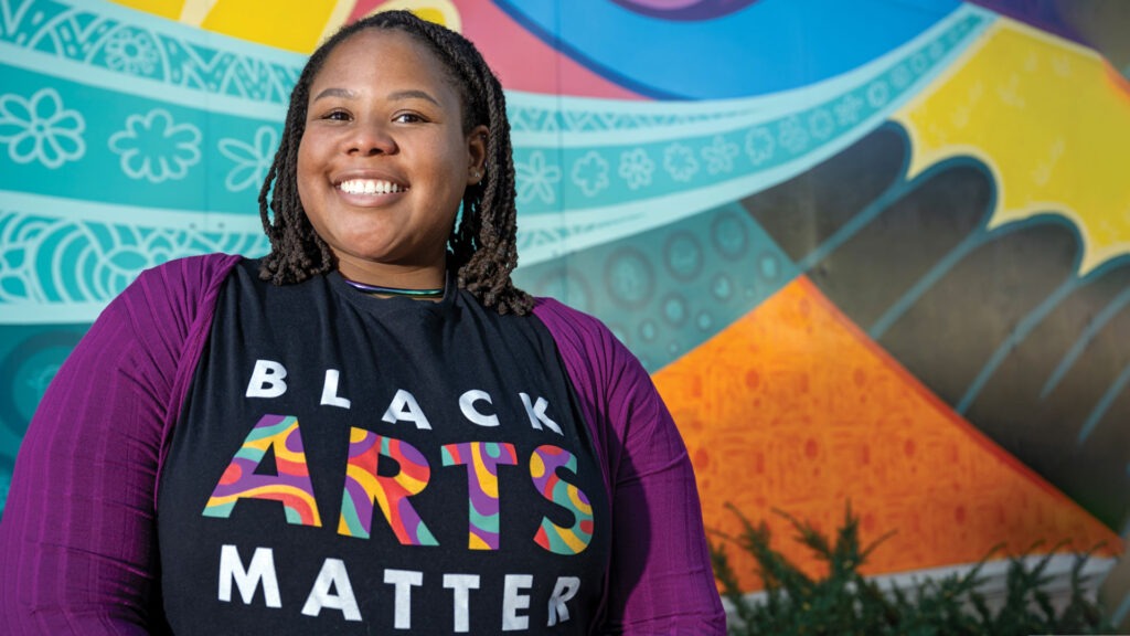 Shasparay Irvin wearing Black Arts Matter t-shirt, standing in front of colorful mural