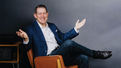 Sean Jacobsohn smiling and shrugging as he sits relaxed in a chair
