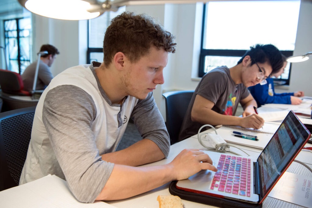A student working on his laptop with other students studying in the background