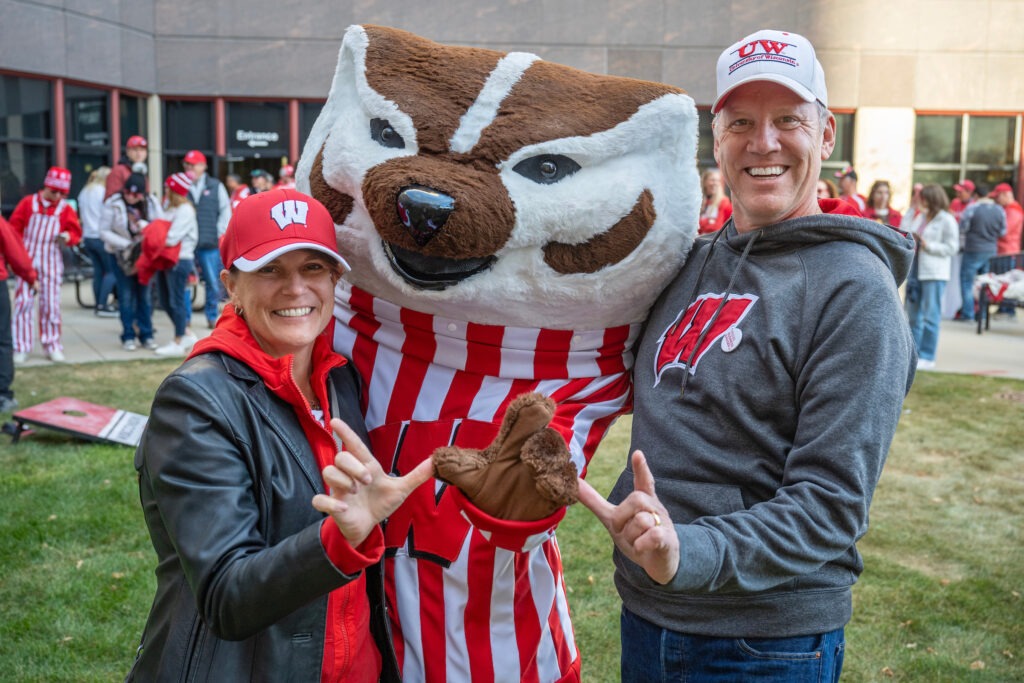 Alumni Chuck Olsen (BA ’87, MBA ’00) and Marjorie Olsen (BS ’96, MBA ’01) smile big as they throw up the "W" with Bucky Badger during WSB's 2024 Homecoming Bash!