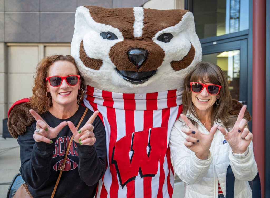 Alum Melissa Siems (BBA ’96) and Aimee Jo Castleberry rock some WSB sunglasses while posing for a photo with Bucky Badger.