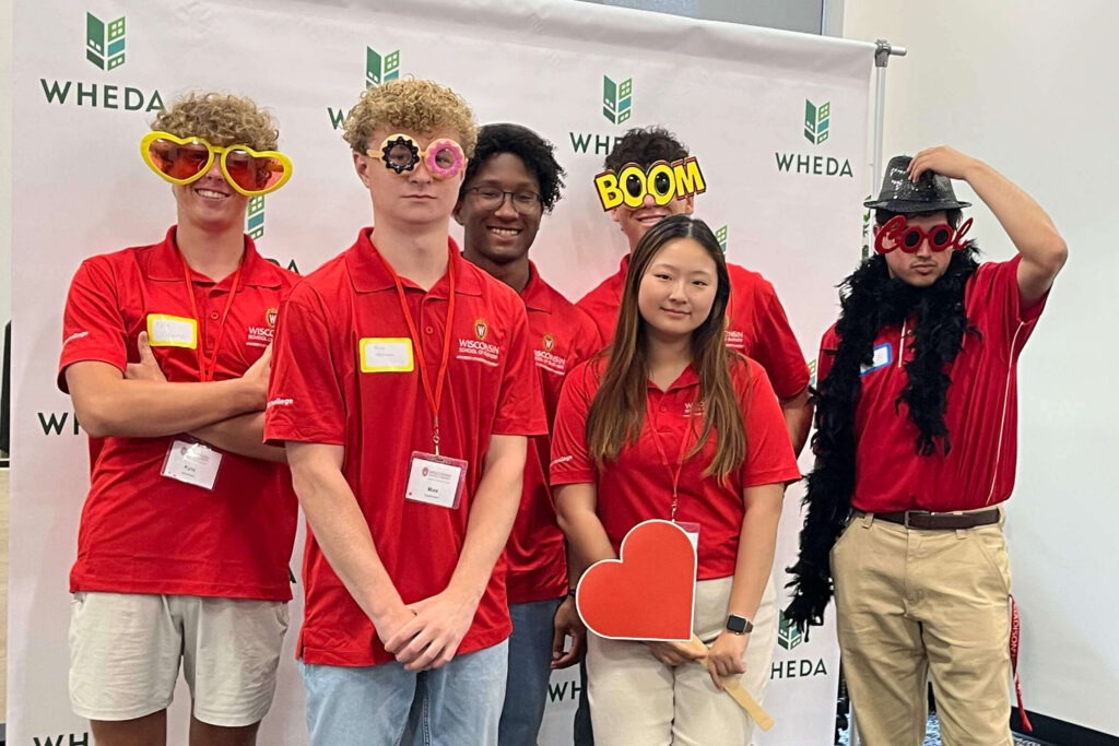 Six students in red shirts standing in front of a WHEDA screen