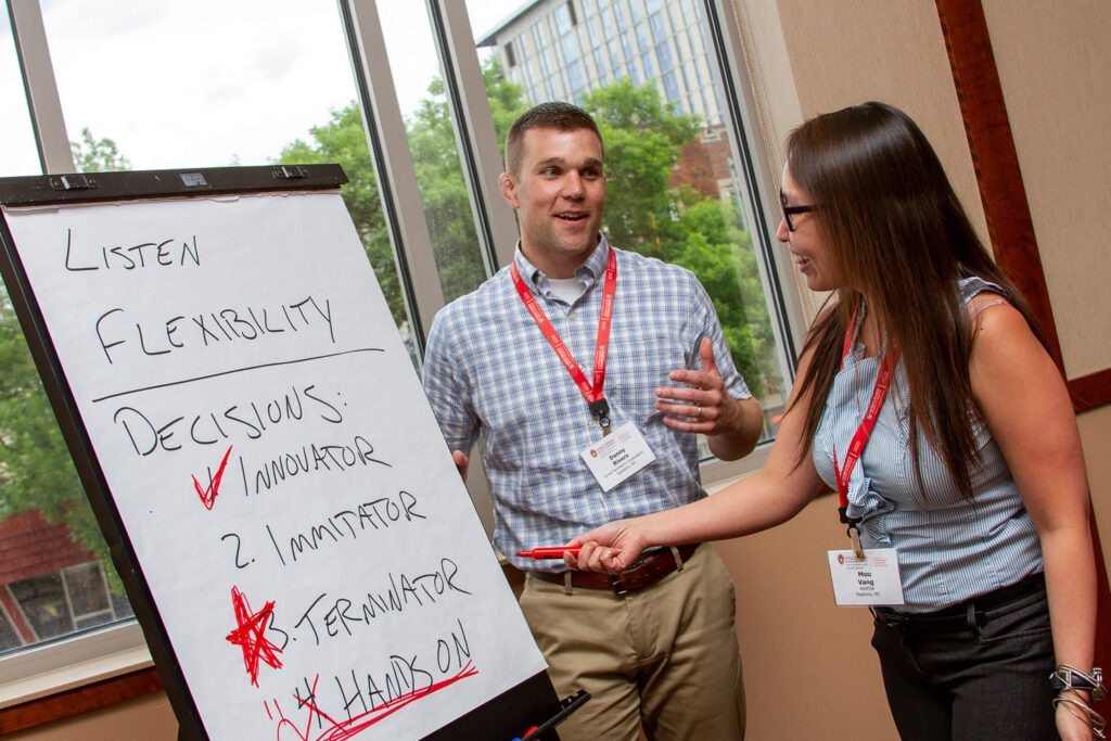 A man and a woman talking in front of a giant white paper pad with the woman pointing at the paper pad with a red marker.