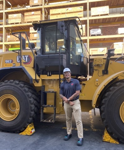 Alexander in a Ferreycorp warehouse, wearing a hardhat, and standing next to a large Caterpiller vehicle