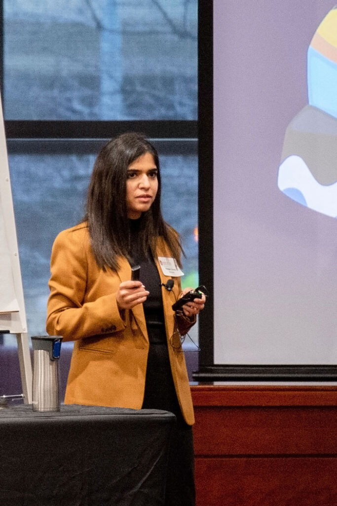 A woman presents to an audience while holding a mic pack and a remote for advancing her slideshow.