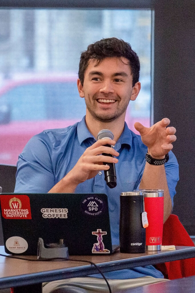 A man sits in front of a table and speaks into a microphone. There is a laptop and coffee thermos on the table.