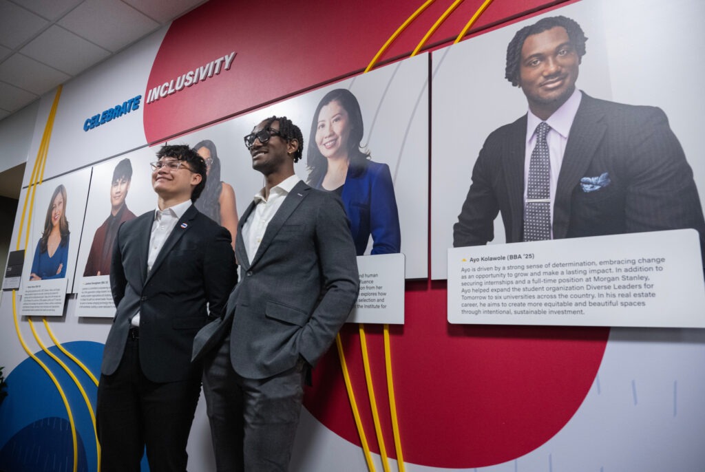 Two students in suits standing in front of wall featuring large them and other notable students and faculty