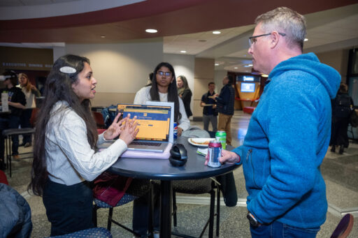 A woman speaks to another woman and a man while standing at a table that holds laptops, food, and drinks.