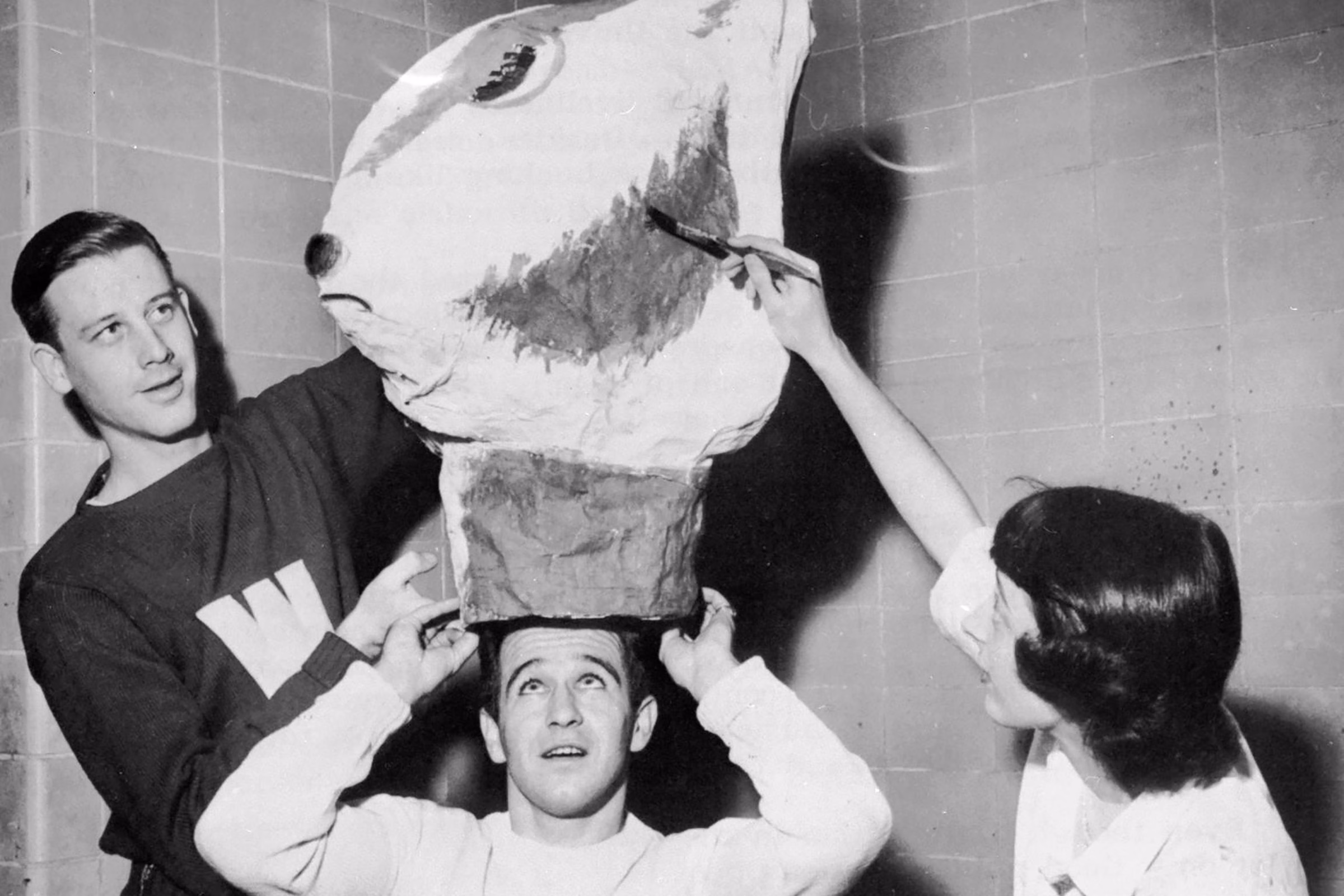 A black and white photograph from a 1949 UW football game showing two students help lift the new Bucky Badger mascot head onto another student's head.