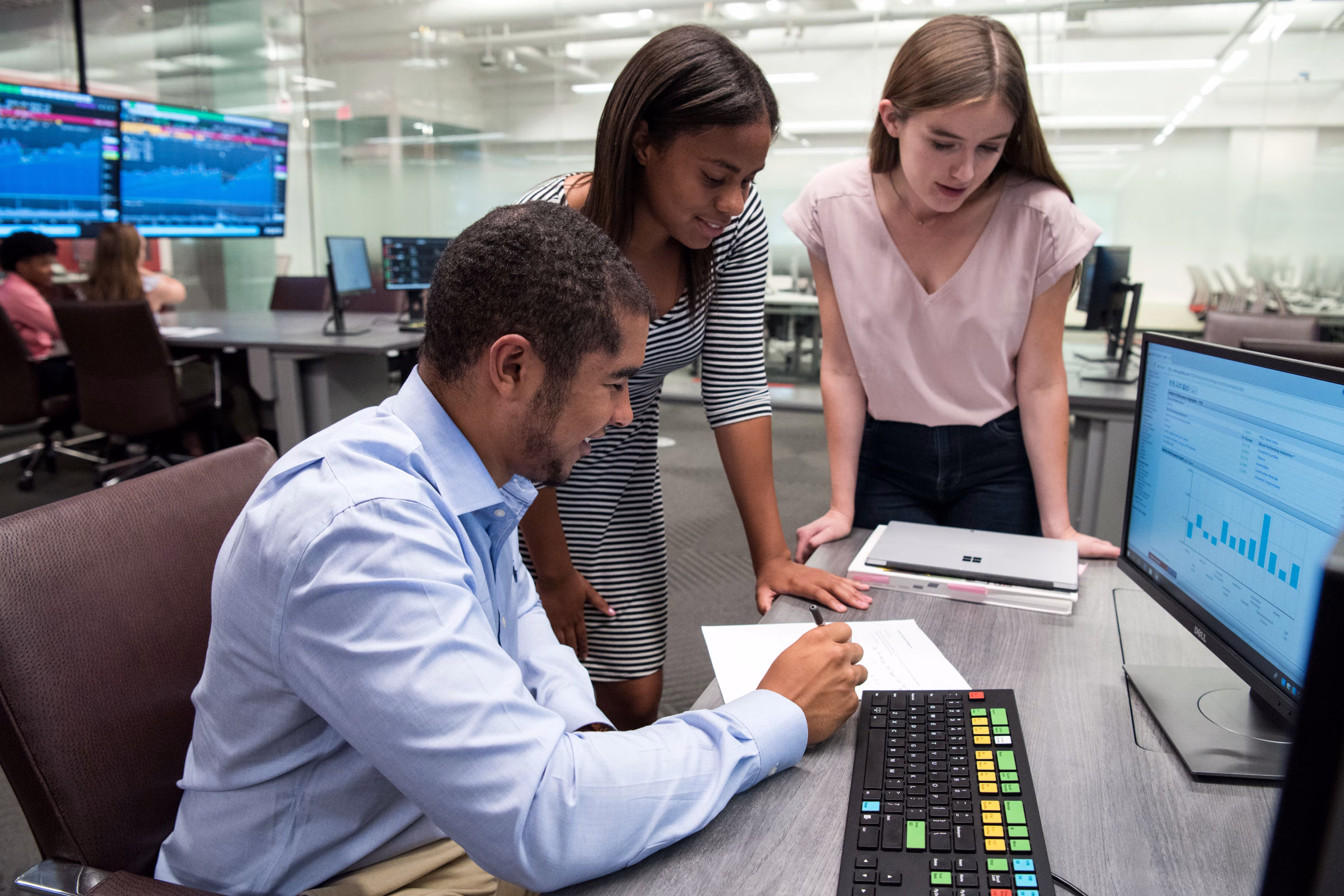 A male student sits in front of a computer while looking at a piece of paper on the table in front of him. Two female students stand nearby, also looking at the paper.