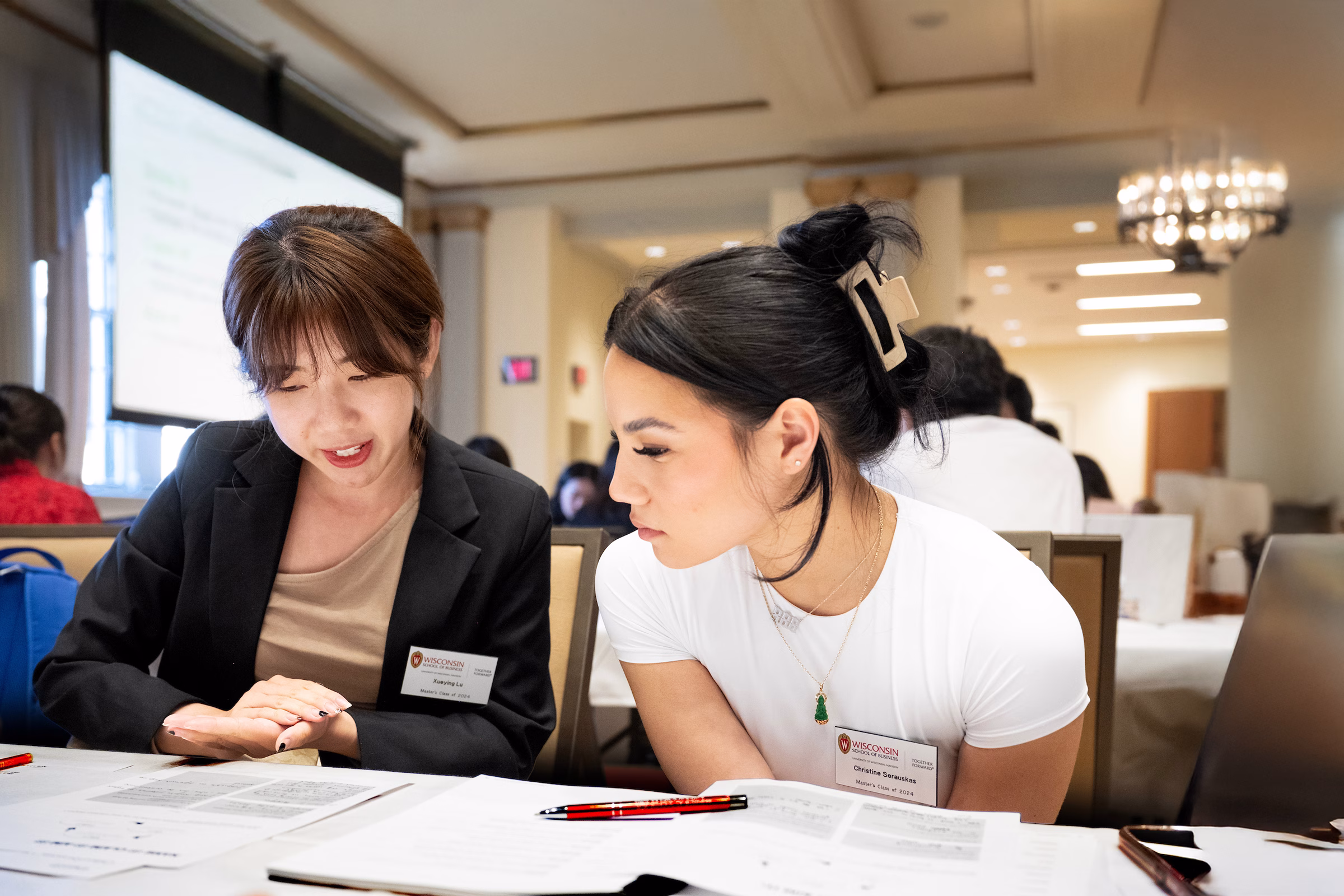 Two women sitting at a table looking at paper on the table.