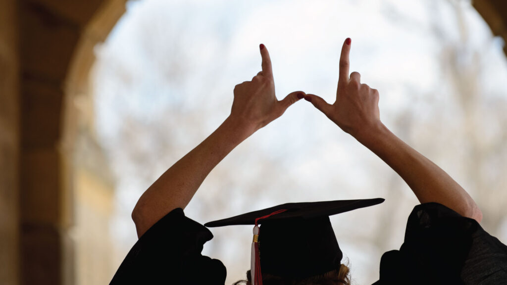 A graduate wearing cap and gown throws a "W" with their hands above their head.