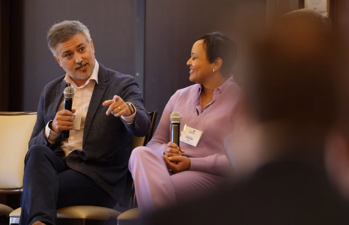 Ben Wright is seated as a panelist and speaking into a mic. A woman is on the right, and the back of a man's head is in the foreground at far right.