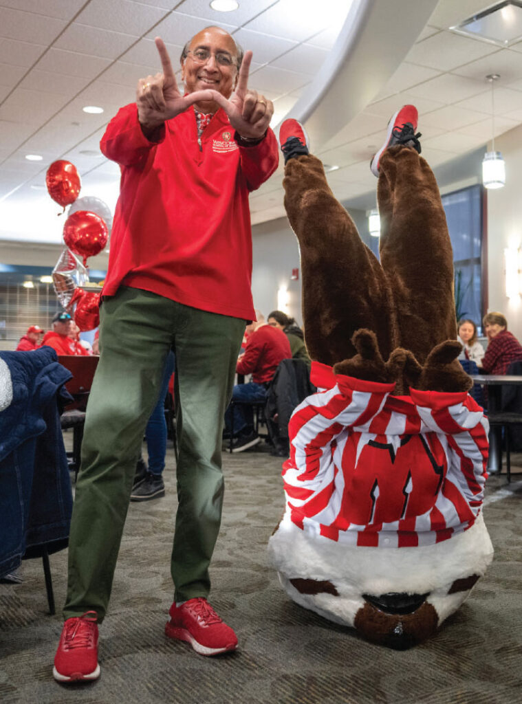 Dean Samba smiles and stands next to Bucky Badger, who is doing a headstand.