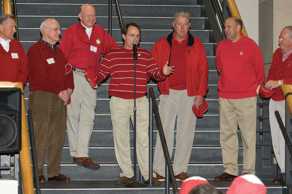 Mike Knetter speaks at a microphone while several men wearing red stand with him.