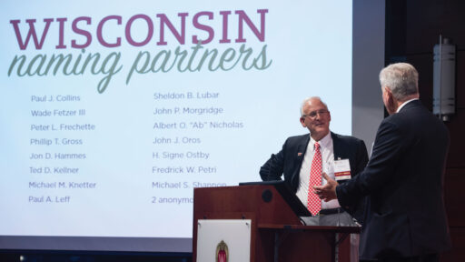 Two men stand on stage and talk about the Wisconsin Naming Partnership.