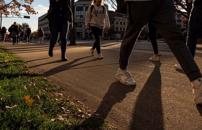 Photo of the legs of students walking on a sidewalk with their legs casting shadows from the early evening sun. Grainger Hall is in the background.