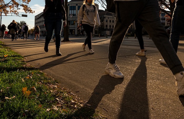 Photo of the legs of students walking on a sidewalk with their legs casting shadows from the early evening sun. Grainger Hall is in the background.