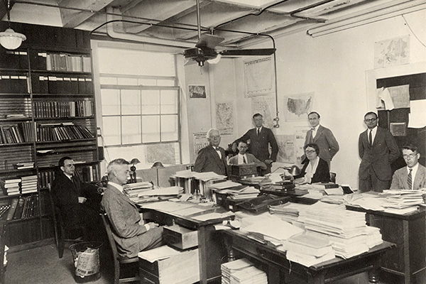 A black and white photo taken inside the School of Commerce in 1929, showing men and women sitting and standing around desks piled with papers.