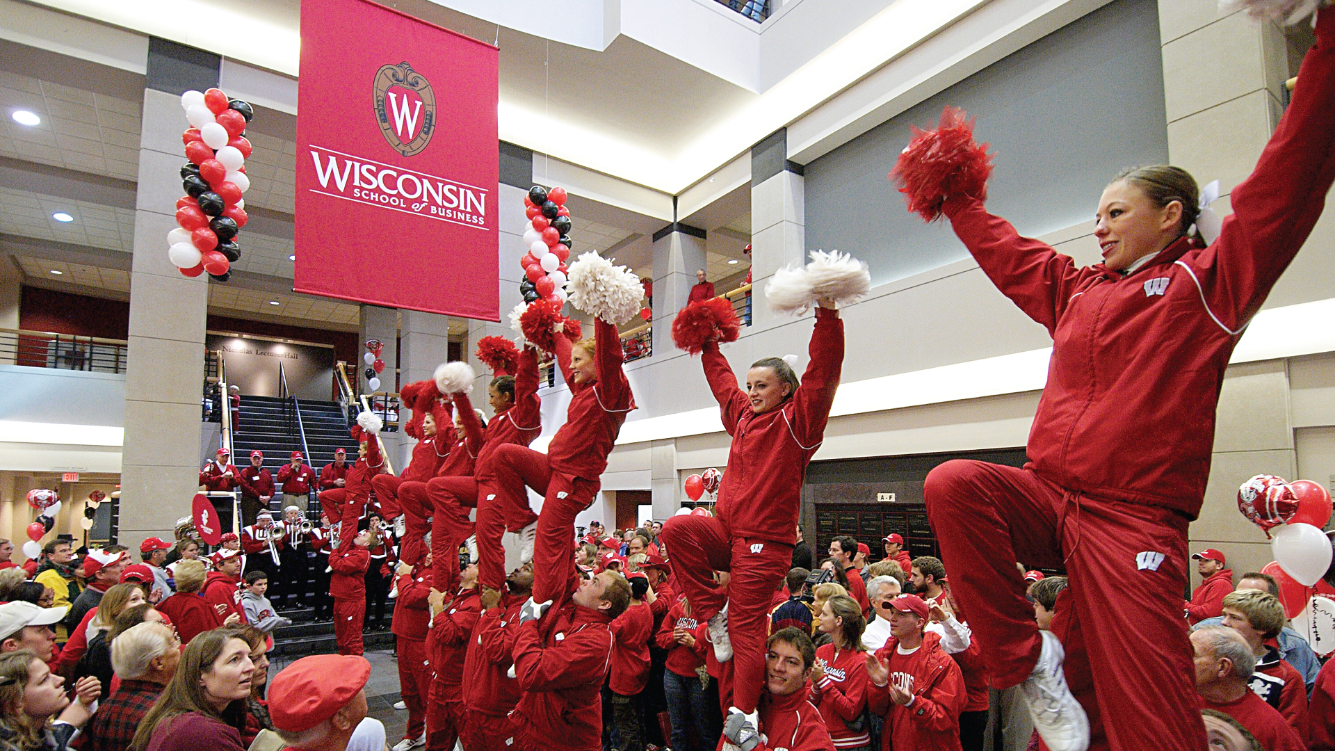 UW marching band members and cheerleaders celebrate homecoming inside Grainger Hall's atrium as red and white balloons fall from the ceiling.