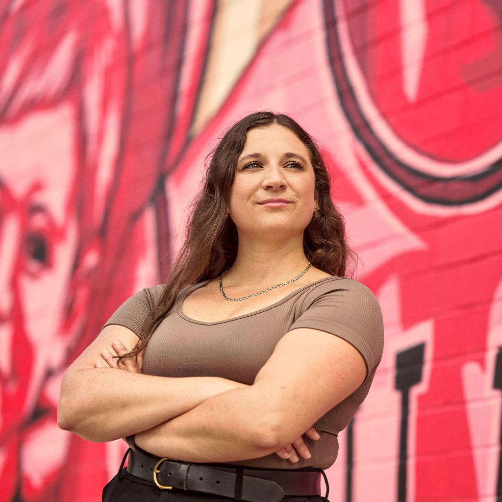 Abbe smiling into the distance with her arms crossed, in front of a mural.