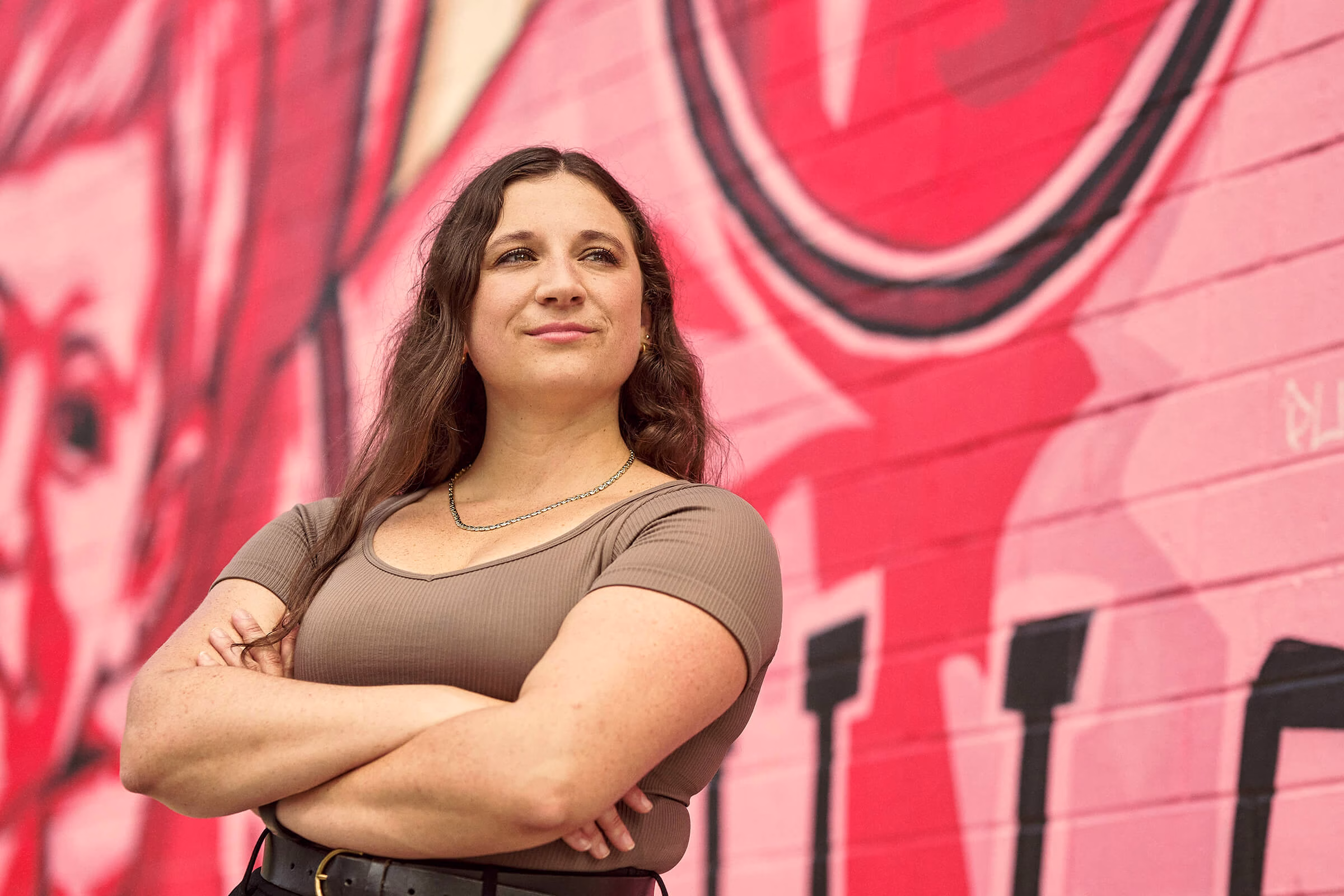Abbe smiling into the distance with her arms crossed, in front of a mural.