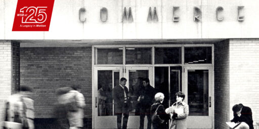 Archival black and white photograph shows the Commerce building with students mingling and talking out in front of it