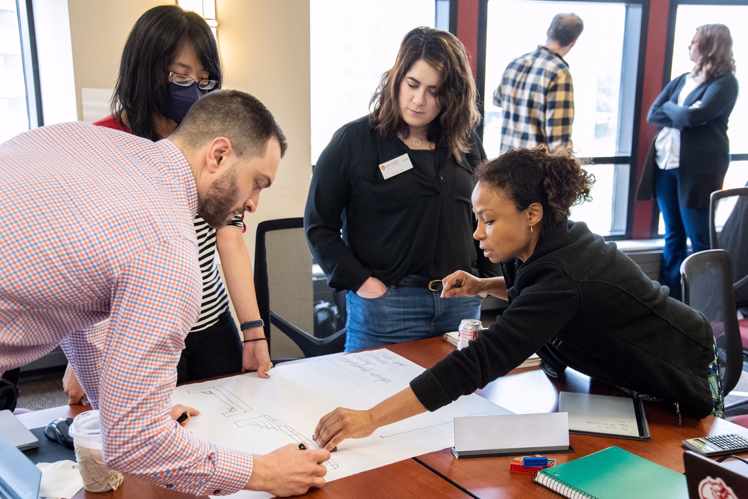 A group of three women and one man are surrounding a desk and making a diagram on a large, white paper.